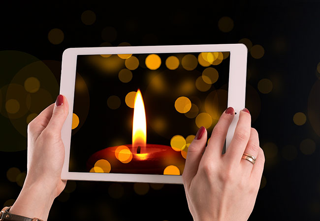 Zu sehen sind zwei Hände, die ein Tablet halten. Darauf zu sehen sind eine Kerze und Lichtpunkte. Bild: pixabay/geralt
